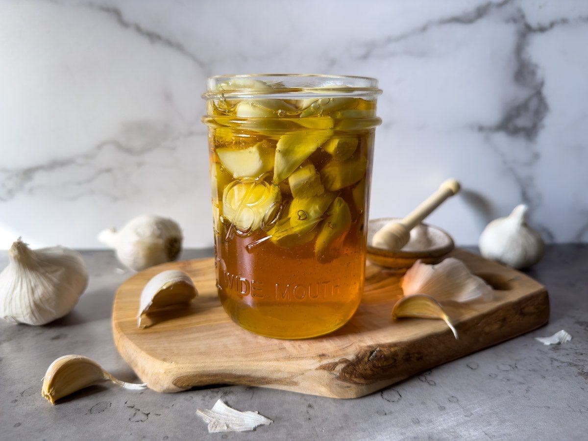 A pint jar full of garlic cloves and honey is sitting on top of a small wood cutting board. Bulbs and cloves of garlic surround the area.