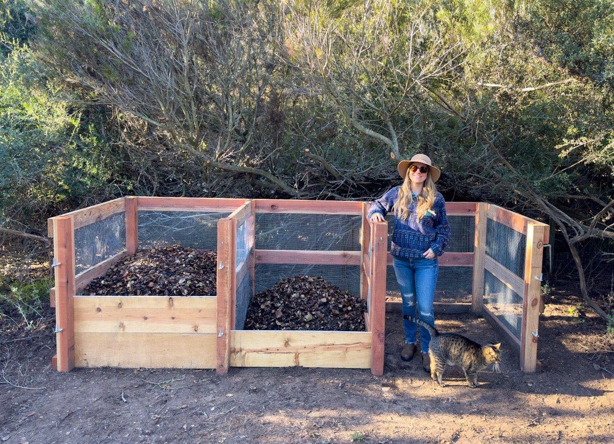 A woman is standing in front of a 3 bay compost system that is made of wood and hardware cloth. Two of the bins are partially filled with compost.