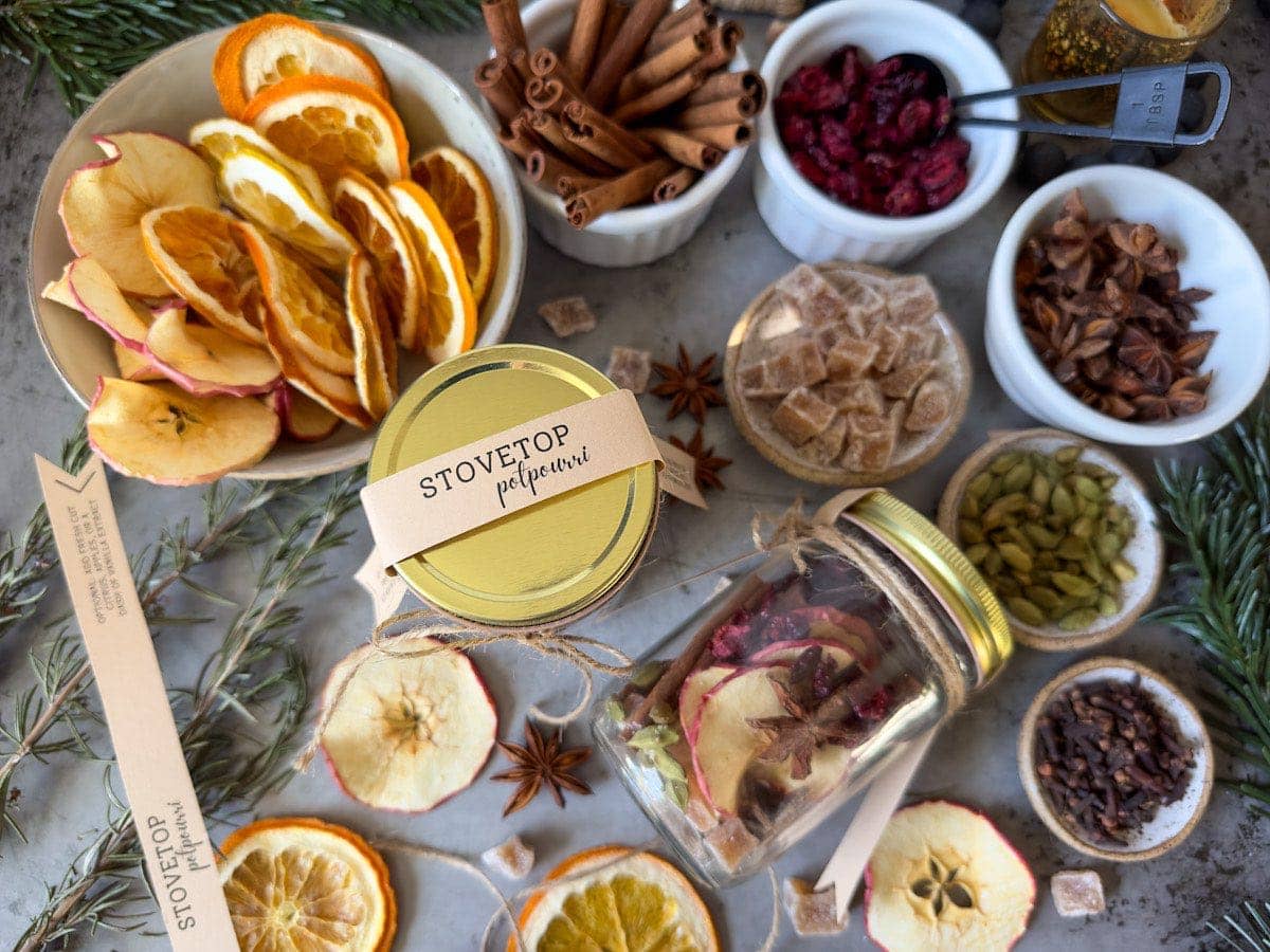 An array of dried orange slices, dried apple slices, ramekins filled with cinnamon sticks, cranberries, star anise, and pinch bowls of crystalized ginger, cardamom, and clove. There are two jars that contain a variety of each of the ingredients for stovetop potpourri.